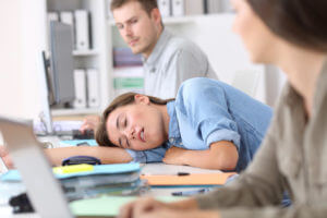 A fatigued female employee is sleeping at the office due to narcolepsy, while her colleagues look at her.